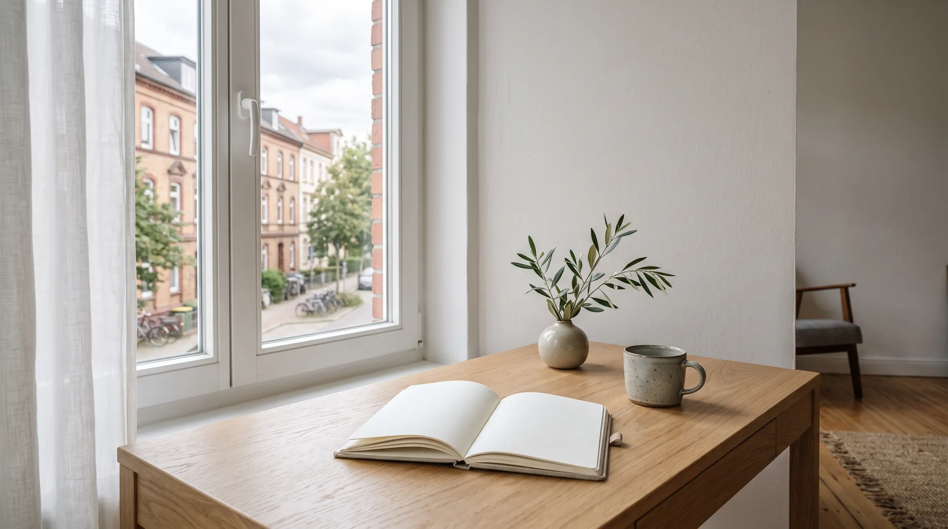 Heller Schreibtisch mit Notizbuch, Tasse und Olivenzweig vor einem Fenster mit Blick auf eine Wohnstraße
